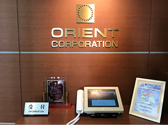 オフィス受付。ゴールドのORIENT CORPORATIONの文字とロゴが正面の壁に掲げられ、テーブルの上には受付用のパネルや盾が飾られている。