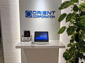オフィス受付。ブルーのORIENT CORPORATIONの文字とロゴが正面の壁に掲げられ、テーブルの上には受付用のパネルや盾が飾られている。