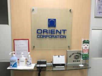 オフィス受付。ORIENT CORPORATIONの文字とロゴが正面の壁に掲げられ、ガラス張りの机の上には受付電話機やカレンダーが置かれている。