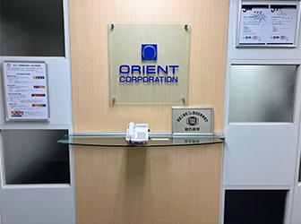 オフィス受付。ORIENT CORPORATIONの文字とロゴが正面の壁に掲げられ、ガラス張りの机の上には受付電話機や盾が置かれている。